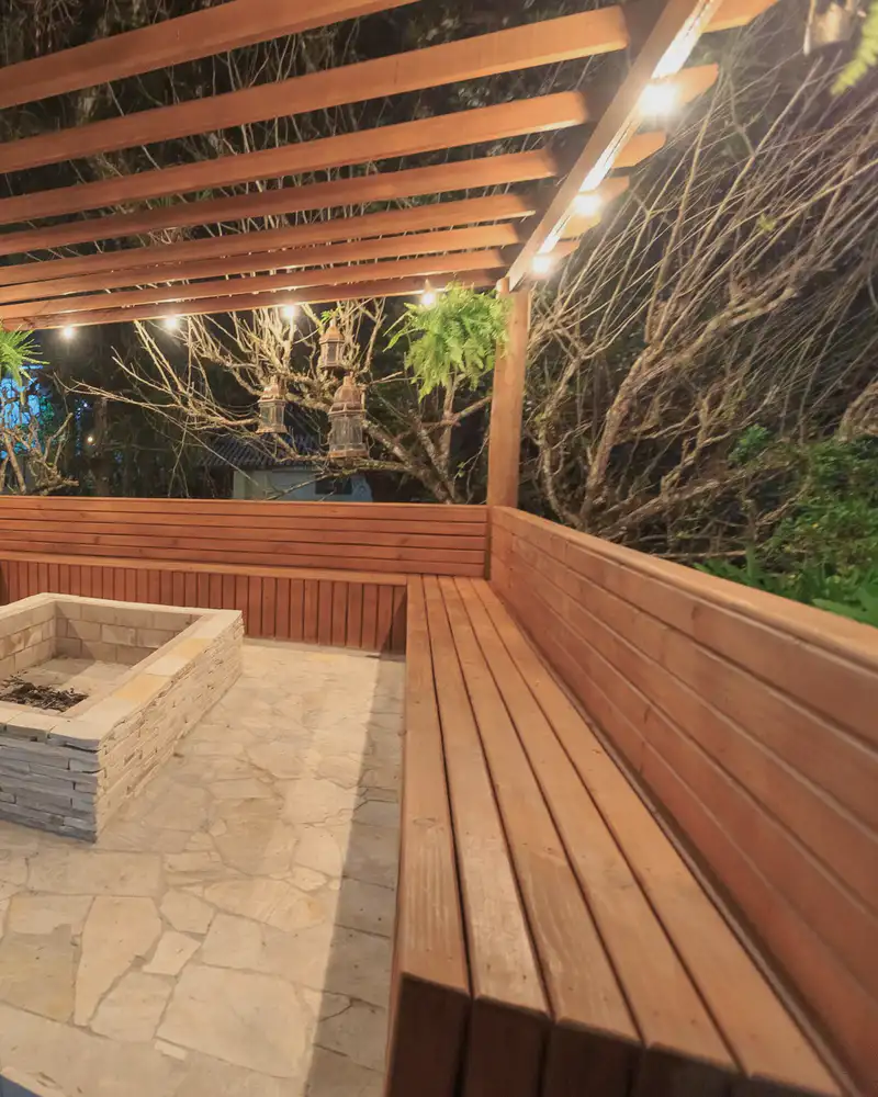 Built-in wood benches around a stone fire pit under a lit pergola